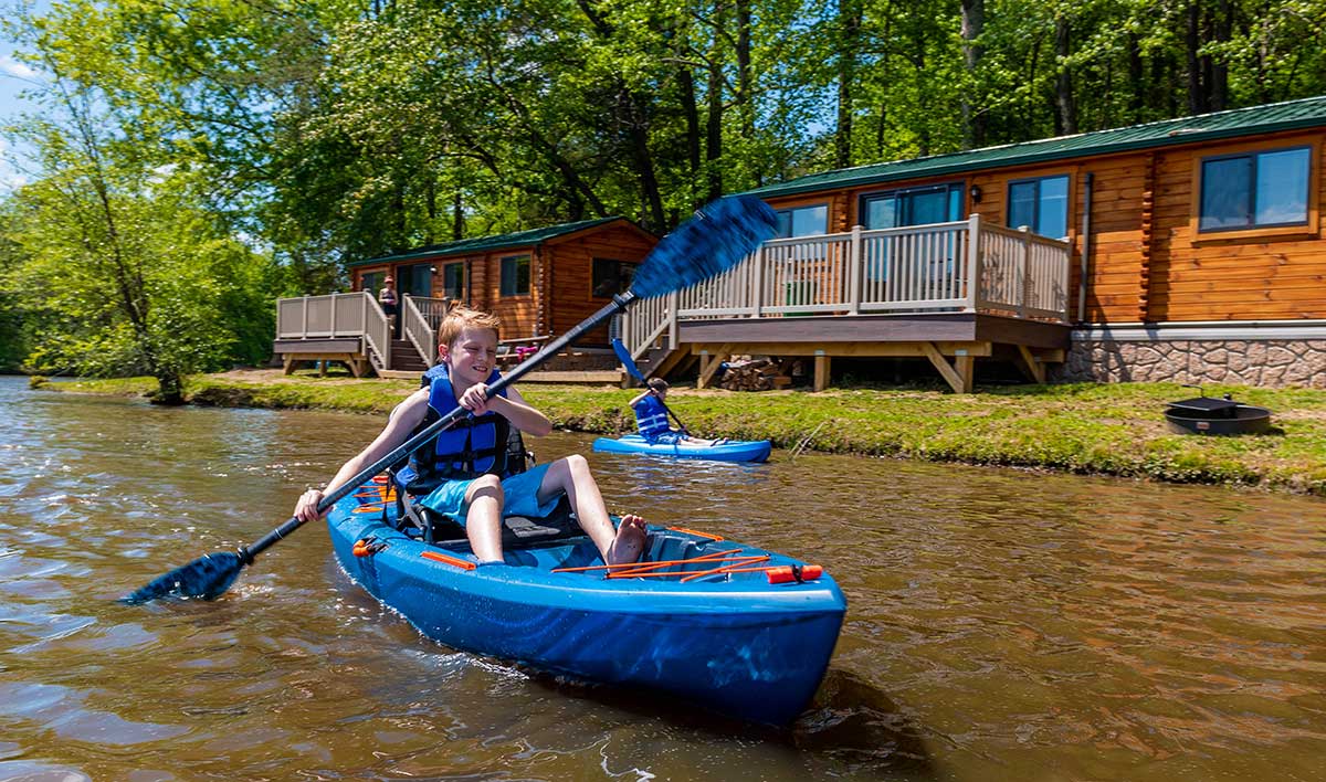 Boys paddling blue kayaks in front of lakeside camp cottages